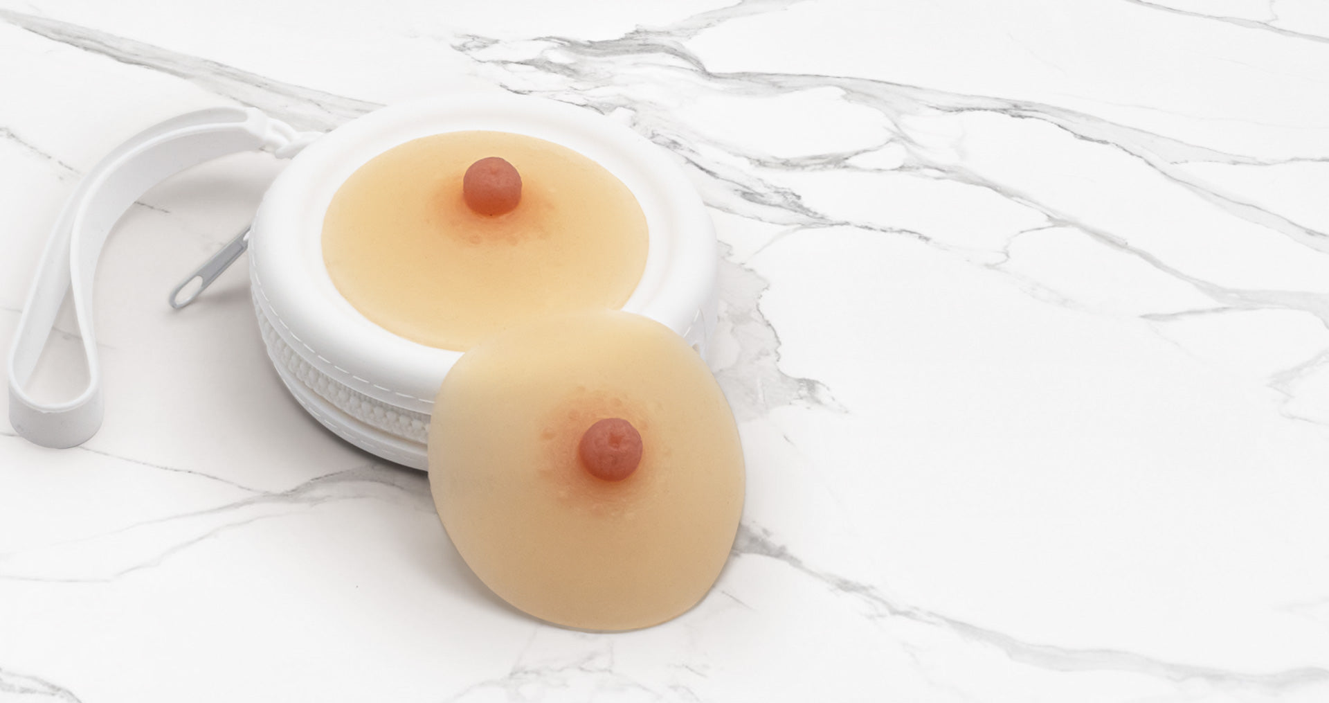 A pair of beige silicone stick-on breast forms with pinkish-red nipples in a white circular case
