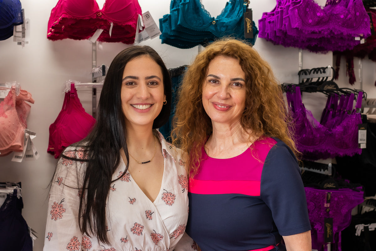 Two women smiling in front of a colorful bra display featuring floral white blouse and navy top with fuchsia stripes