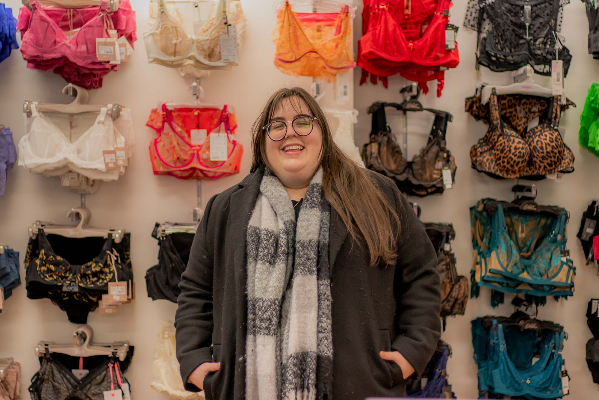 A smiling woman with long brown hair and glasses stands in front of colorful bras wearing a dark coat and patterned scarf