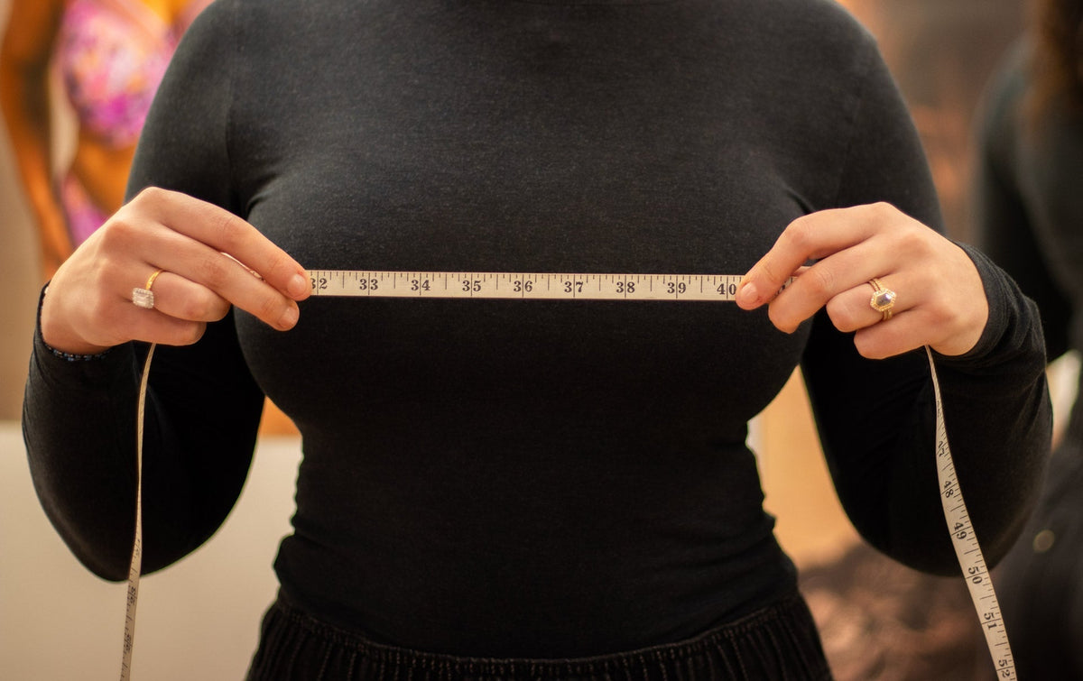 A white flexible measuring tape with black numbers held taut across a person’s chest illustrating bra size measurement.