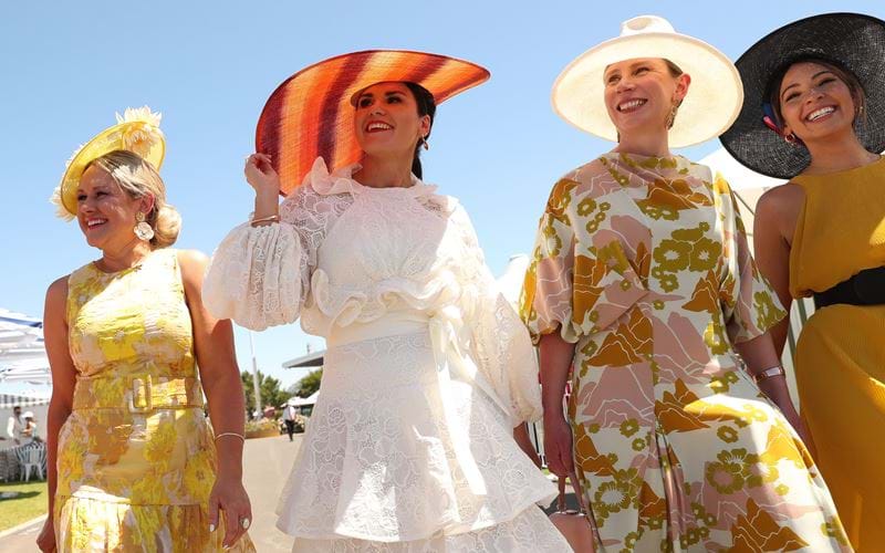 A woman wearing a white lace dress with puffed sleeves and an orange red striped wide brimmed hat off to the races