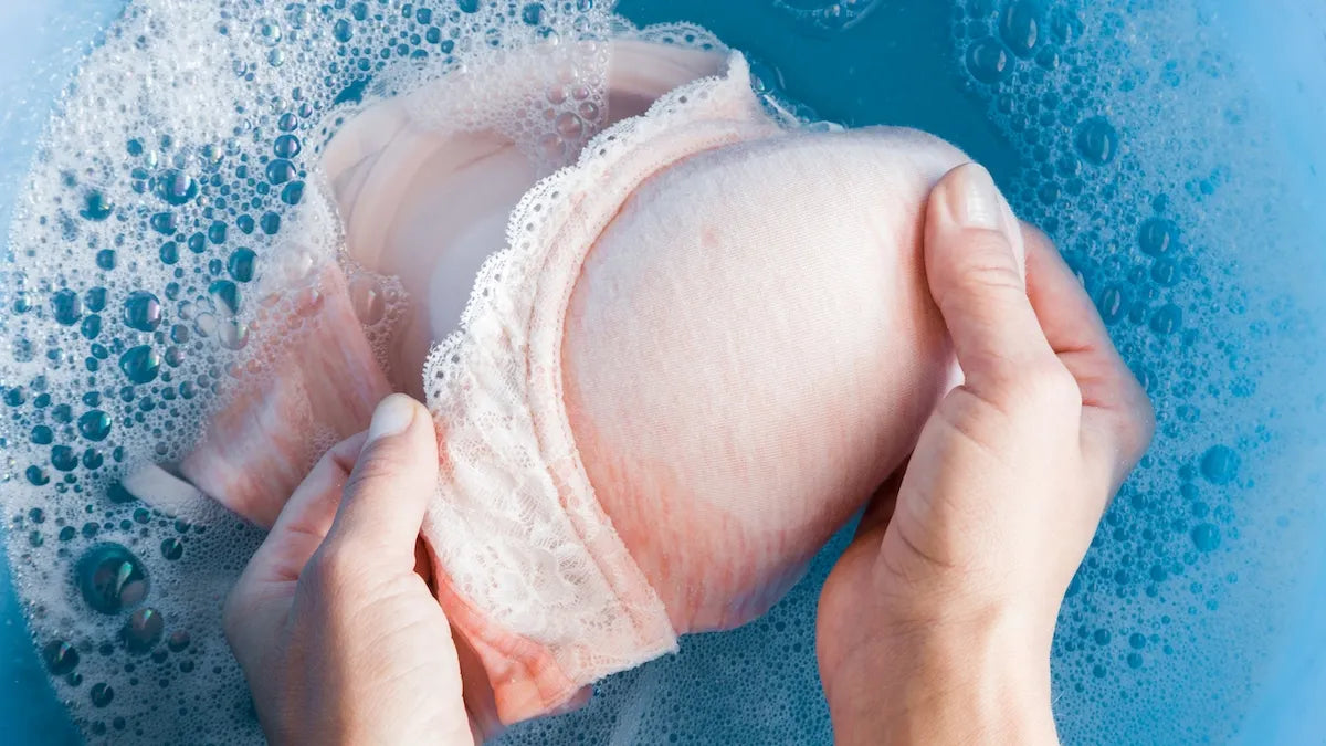 hands washing a pink bra in blue soapy water