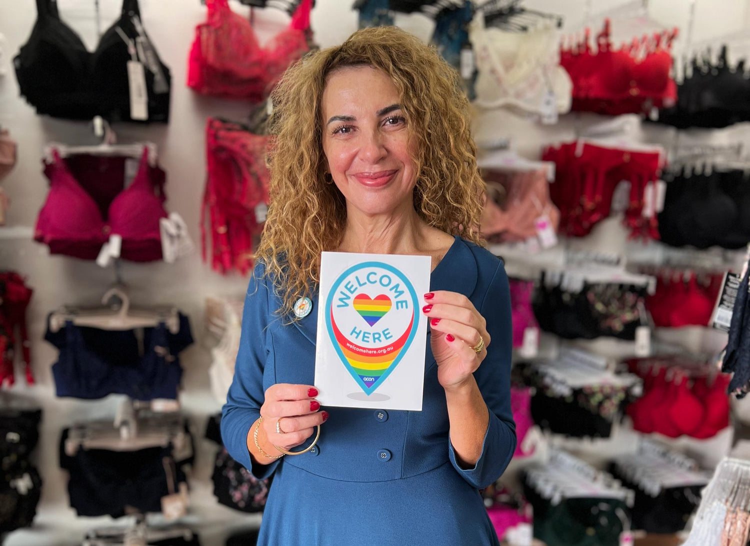 A woman holding a white rectangular sign with a rainbow heart and blue welcome here text in an article titled Everyone is Welcome Here