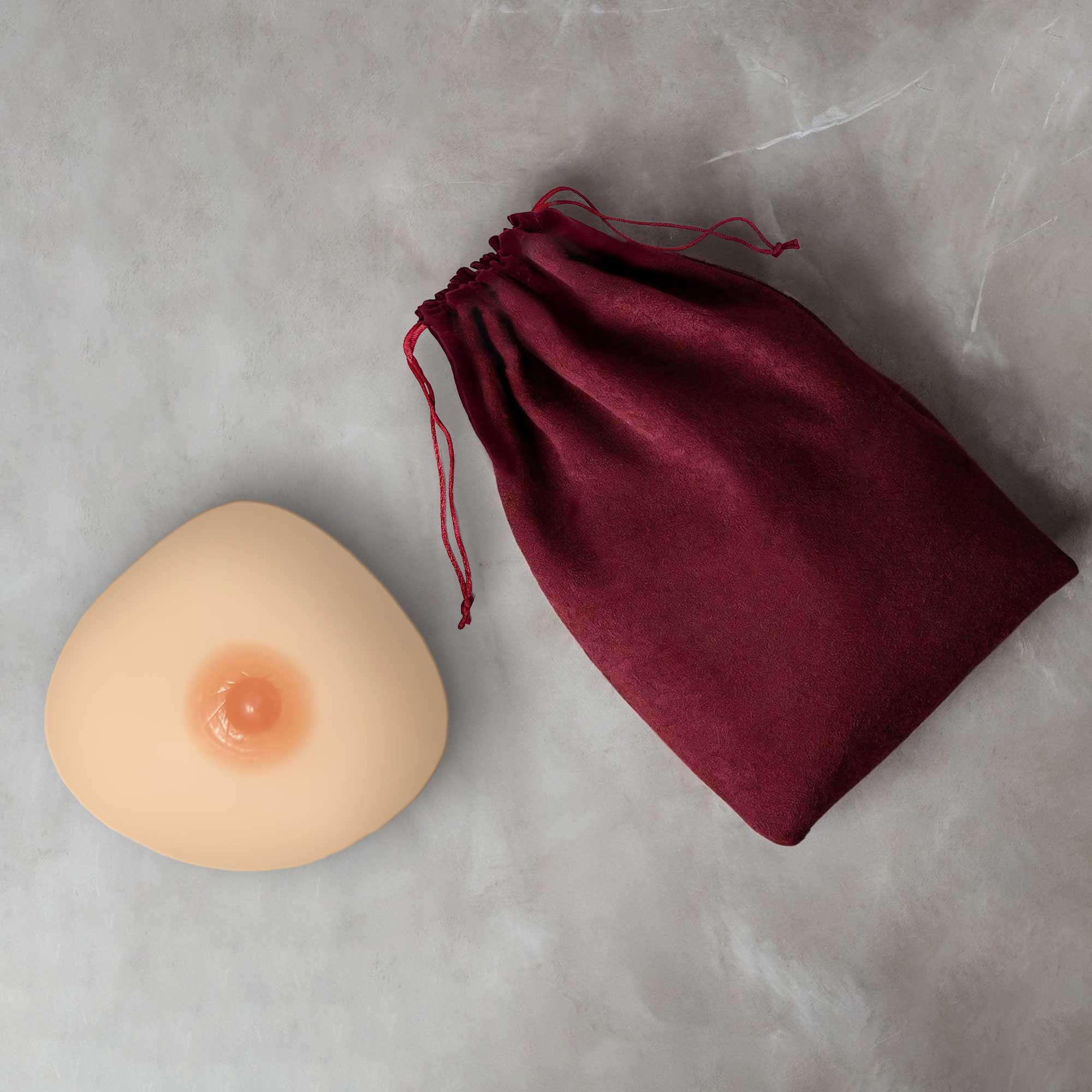 Beige silicone breast form with natural pink nipple beside burgundy drawstring bag from illusions lingerie collection