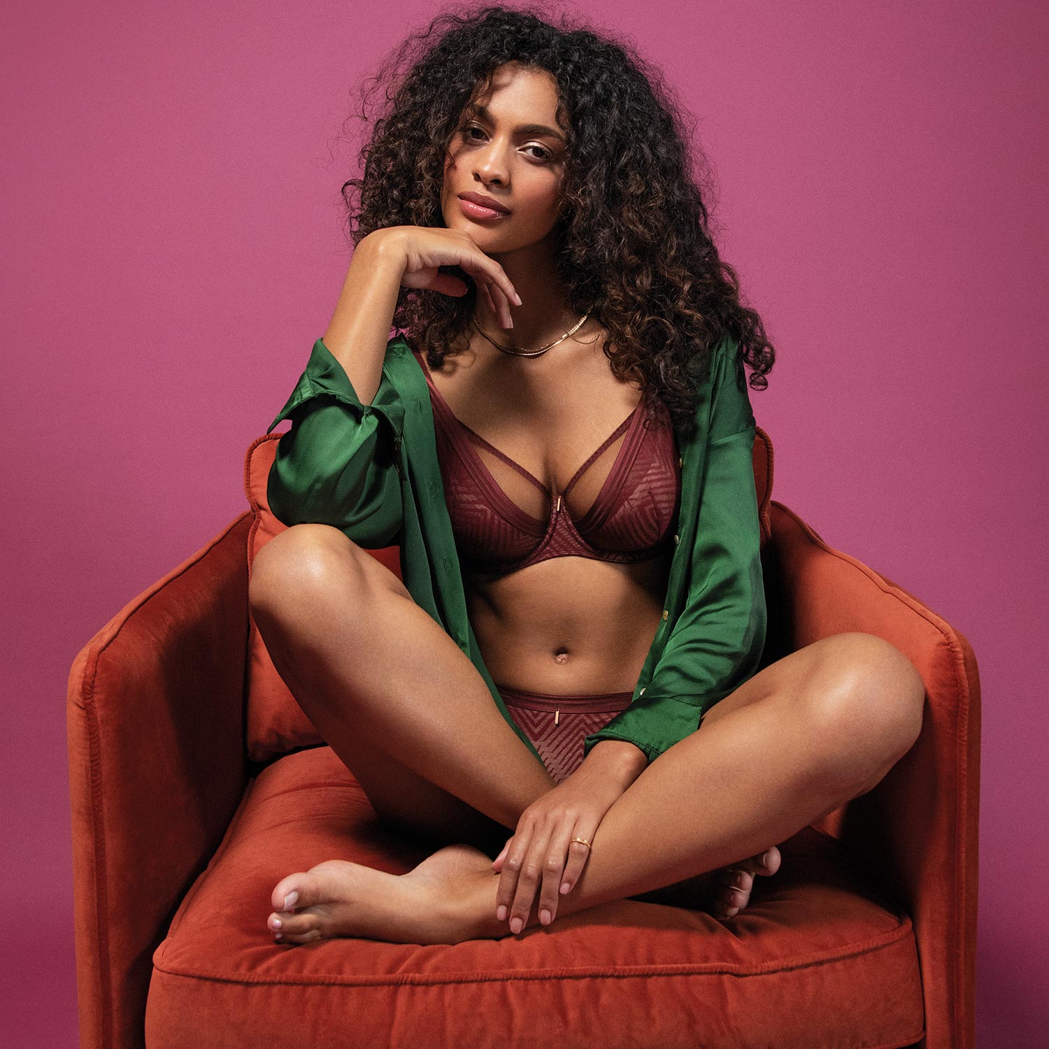 The model is wearing a maroon lace bra with geometric patterns and matching briefs with an open green satin robe from Freya.