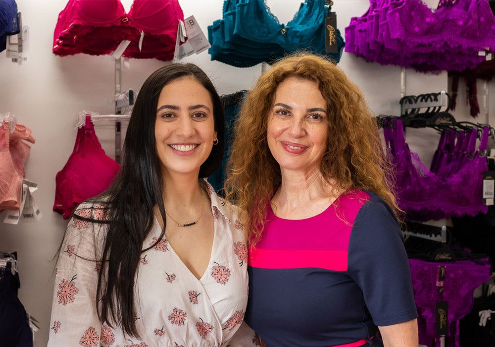 Two bra fitters standing side by side in front of a wall of colourful bras