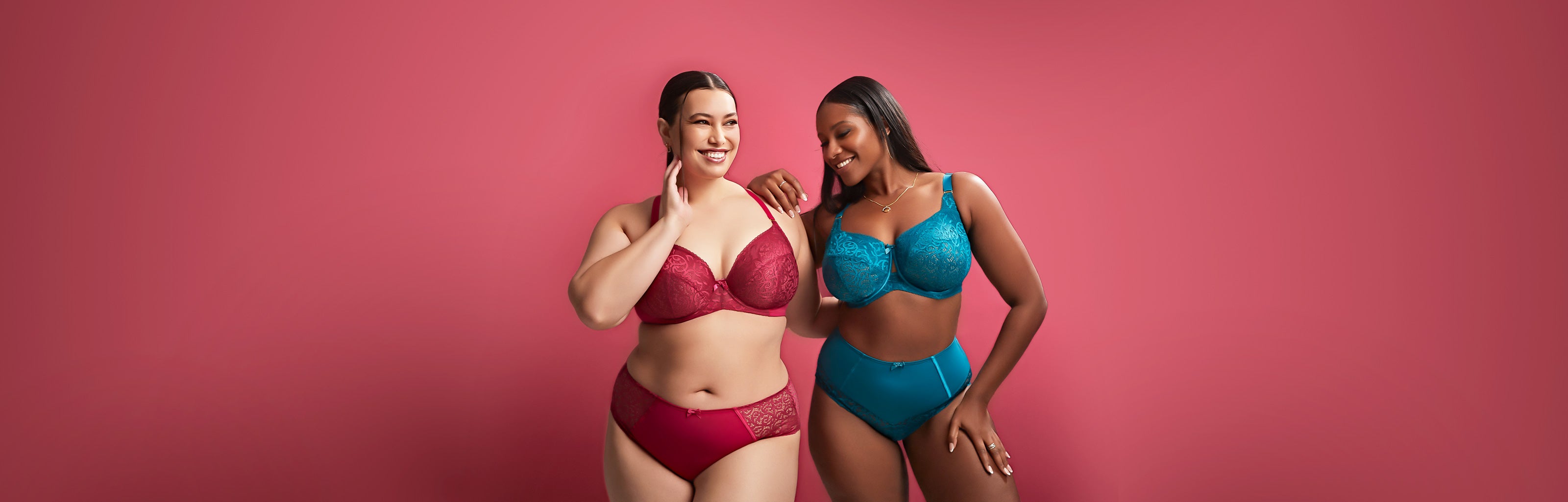 Two full figure women in colourful lingerie smiling after a bra fitting on a light red background