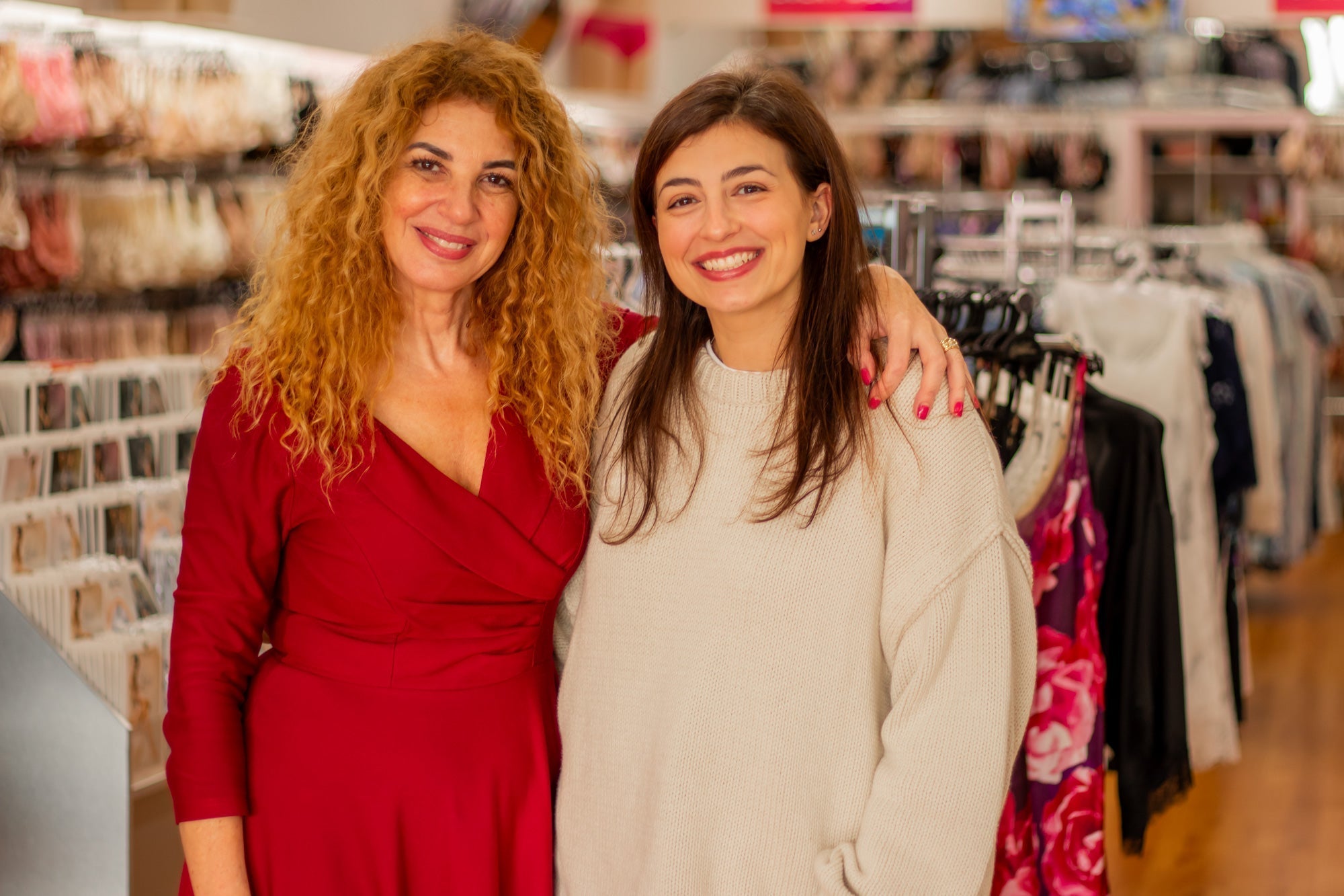 The woman on the left wears a vibrant red long-sleeved wrap dress with a flattering v-neckline in Meet Our Newest Bra Fitter Angelique